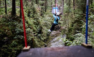Ein Kind schwingt an einer Seilrutsche zwischen Bäumen in einem grünen Wald. Umgeben von Natur und einem schmalen Bach darunter. | © Kleinwalsertal Tourismus | Oliver Farys