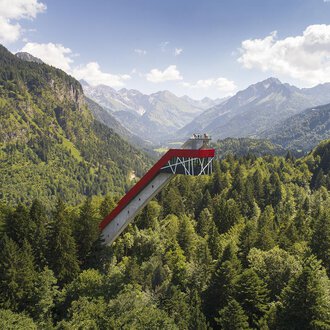 Eine beeindruckende Aussichtsplattform, die über die Bäume ragt, umgeben von majestätischen Bergen und dichten Wäldern. Der Himmel ist klar und zeigt einige Wolken.