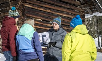 A group of five people stands in the snow in front of a wooden barn. They are collectively looking at a photo that one of the participants is holding in their hand. | © Kleinwalsertal Tourismus | Dominik Berchtold