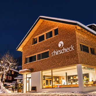 A modern building with a wooden facade and large windows, illuminated at night. It is surrounded by snow and located in a picturesque winter setting.
