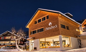 A modern building with a wooden facade and large windows, illuminated at night. It is surrounded by snow and located in a picturesque winter setting.