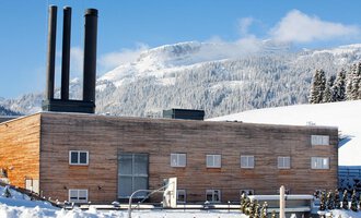 A modern building with a wooden facade and three chimneys in a snow-covered landscape. In the background, mountains and a clear blue sky can be seen.
