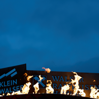 Ein Werbeschild mit der Aufschrift "Klein Walser" brennt im Dunkeln. Flammen leuchten gegen den blauen Himmel. | © Kleinwalsertal Tourismus eGen | Oliver Farys