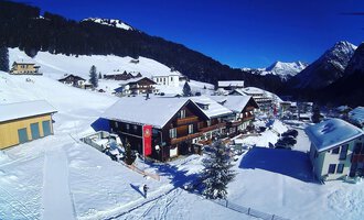 Eine schneebedeckte Landschaft mit gemütlichen Holzhaus und Bergen im Hintergrund. Der Himmel ist klar und blau.