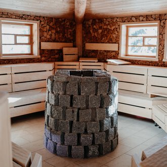 Ein gemütlicher Saunaraum mit Holzbänken und großen Fenstern. In der Mitte steht ein Ofen aus Steinen. | © Verwöhn- & Wellnesshotel Walserhof| Aileen Melucci
