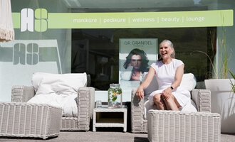 A relaxed woman is sitting in a modern lounge area with woven furniture. In the background, wellness and beauty elements are visible. | © Annett Schwärzler Nails & Spa | Stefanie Haid