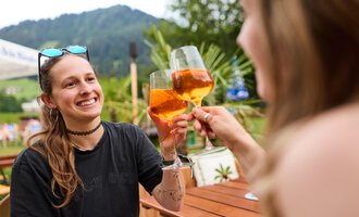 Two women are toasting with drinks and smiling. In the background, green mountains and plants can be seen. | © Kleinwalsertal Tourismus | Oliver Farys