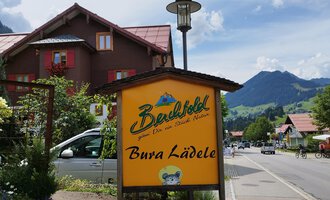 Ein rustikales Schild von "Bergblick" mit der Aufschrift "Bura Lädele". Im Hintergrund sind typische Alpenhäuser und Berge zu sehen. | © Kleinwalsertal Tourismus | N. Lughammer