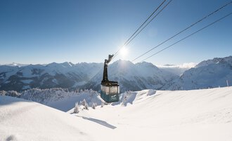 Eine Bergbahn schwebt über eine schneebedeckte Landschaft. Im Hintergrund erstrecken sich majestätische Berge unter einem klaren blauen Himmel. | © OBERSTDORF · KLEINWALSERTAL BERGBAHNEN
