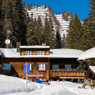 A cozy mountain cabin in winter with a snow-covered ground. In the background, high mountains and green fir trees can be seen. | © Bühlalpe | Frank Drechsel