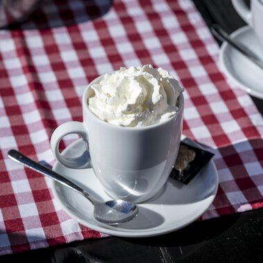 A cup with cream on a white saucer. The background is a red and white checked table runner. | © Kleinwalsertal Tourismus | Dominik Berchtold