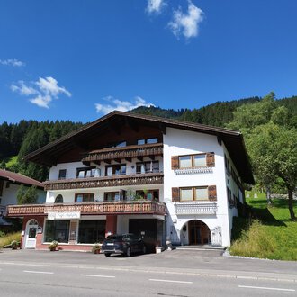A charming building in alpine style with various balconies. In the background, green hills and a blue sky can be seen. | © Kleinwalsertal Tourismus | Nicole Lughammer