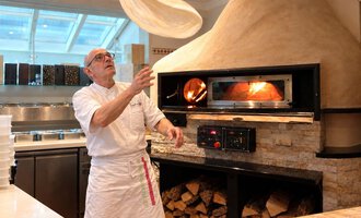 A chef throws a pizza dough into the air. In the background, there is a traditional stone oven.