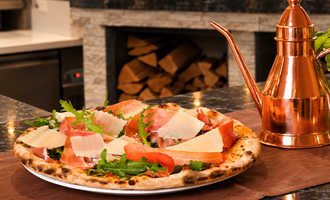 Eine leckere Pizza mit Schinken, Parmesan und frischem Rucola. Im Hintergrund steht eine copperfarbene Ölpresse.