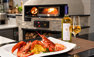 Ein stilvolles Gericht mit Hummer und Pasta auf einem weißen Teller. Im Hintergrund befindet sich ein Ofen und ein Glas Weißwein steht daneben.