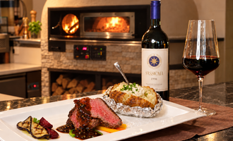 Ein hochwertiges Gericht mit saftigem Steak, einer gefüllten Kartoffel und buntem Gemüse. Im Hintergrund steht eine Flasche Wein und ein Holzofen ist sichtbar.