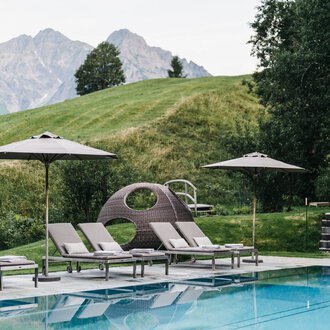 Ein Poolbereich mit Liegen und Sonnenschirmen. Im Hintergrund sind sanfte Hügel und Berge zu sehen.