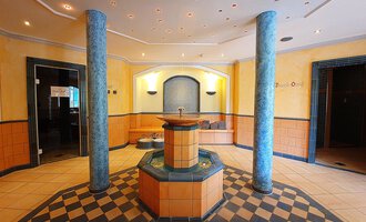 Ein eleganter Wellnessbereich mit einem zentralen Brunnen. Die Wände sind in warmen Farben gestaltet, und der Boden hat ein auffälliges Schachbrettmuster. | © Hotel Birkenhöhe