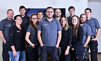 A group of twelve people stands together and smiles. They are wearing similar gray work clothing and are in a bright room. | © Der Elektriker Markus Leitgeb GmbH