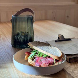 A sandwich with roast beef and arugula on a plate. In the background, there is a green lantern on a wooden table. | © Einkehr Guntschau
