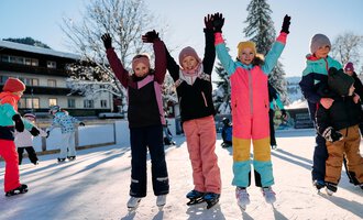 Eine Gruppe fröhlicher Kinder steht auf einer Eislauffläche und hebt die Hände in die Luft. Sie tragen bunte Winterkleidung und genießen den sonnigen Wintertag. | © Kleinwalsertal Tourismus | Oliver Farys