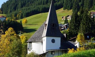 Eine malerische Kirche mit einem hohen spitzen Turm steht in einer grünen Landschaft. Im Hintergrund sind sanfte Hügel und einige Häuser zu sehen. | © Ev. Kirche Kleinwalsertal