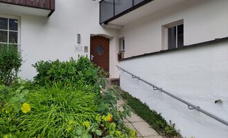 An entrance area of a house with a green, blooming garden landscape. The front door is made of wood and has a unique shape. | © Ev. Kirche Kleinwalsertal