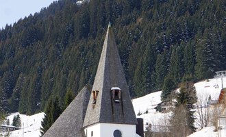 A charming church with a pointed tower, surrounded by snow-covered meadows and wooded mountains. The sky is clear and the landscape appears wintery and peaceful. | © Ev. Kirche Kleinwalsertal