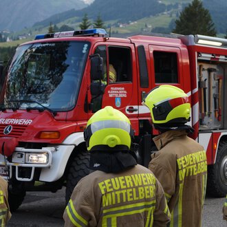 A fire truck is standing on a road, surrounded by firefighters in uniform yellow helmets. The landscape in the background shows trees and mountains. | © Feuerwehr Mittelberg | Christoph Hilbrand