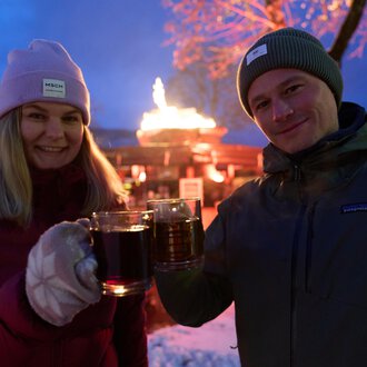 Zwei Personen stoßen mit Gläsern an und lächeln. Im Hintergrund ist eine festliche Atmosphäre mit einer Feuerstelle und 겨우nlichen Lichtern zu sehen.