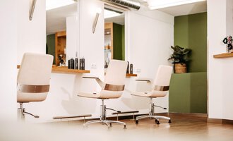 Ein moderner Friseursalon mit eleganten, weißen Friseurstühlen und großen Spiegeln. Die Wände sind in neutralen Farben gehalten, mit einem Hauch von grünem Akzent. | © Friseur Haarwerkstatt | Franziska Diener