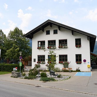 A charming community building with blooming flower boxes. In the background, additional buildings and trees can be seen. | © Kleinwalsertal Tourismus | Oliver Farys