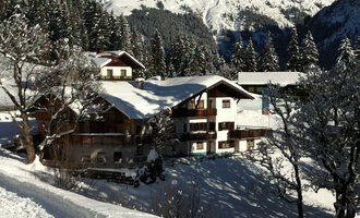 Eine malerische Winterlandschaft mit schneebedeckten Bergen im Hintergrund. Im Vordergrund steht ein charmantes Haus, umgeben von schneebedeckten Bäumen. | © Gasthof Café Alpenwald | Thorsten Alpenwald