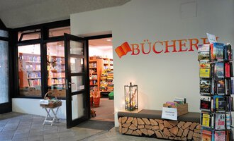 Ein einladender Eingang zu einer Bücherei mit einer großen Auswahl an Büchern im Inneren. Daneben steht ein Bücherregal und eine kleine Sitzgelegenheit. | © Kleinwalsertal Tourismus | S. Guggenberger