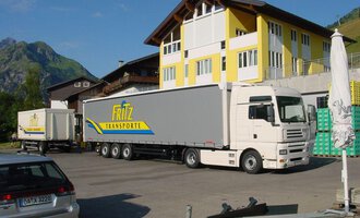 A truck from Fritz Transporte is parked in front of a large building. In the background, green mountains can be seen. | © Getränke Service Fritz | Helmut Fritz