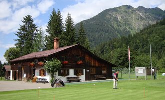 A traditional wooden house surrounded by mountains and trees. In the foreground, there is a well-maintained golf course. | © Golfclub Oberstdorf | ©kunst.joachimweiler.de