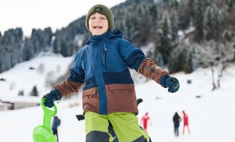 Ein fröhlicher Junge trägt eine bunte Winterjacke und hält einen Schlitten in der Hand. Im Hintergrund sieht man schneebedeckte Landschaften und andere Menschen im Schnee. | © Kleinwalsertal Tourismus | Oliver Farys