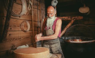Ein älterer Mann mit langen weißen Haaren und Bart arbeitet in einer Holzwerkstatt. Im Hintergrund ist ein großer Kessel zu sehen. | © Käsestadl Kleinwalsertal | Nadine Paul