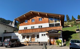 Ein Holzgebäude mit einem Geschäft namens "KäseStadl". Vor dem Haus stehen mehrere Autos, und im Hintergrund sind grüne Hügel zu sehen. | © Käsestadl Kleinwalsertal | Nadine Paul