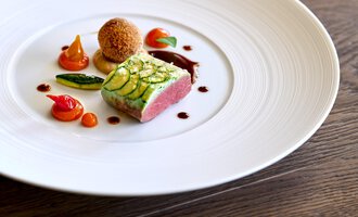 Ein stilvolles Gericht auf einem weißen Teller, bestehend aus Fleisch mit Zucchini, umgeben von bunten Gemüsesorten und einer Sauce. Die Präsentation ist elegant und ansprechend.