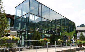 Ein modernes Gebäude mit Glasfront, umgeben von Bäumen und Blumen. Im Vordergrund ist ein Gehweg zu sehen, auf dem Menschen gehen. | © Kleinwalsertal Tourismus | Oliver Farys