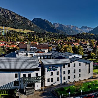 A modern building surrounded by mountains and trees. The sky is clear and the surroundings feel calm and inviting. | © Klinikverbund Allgäu