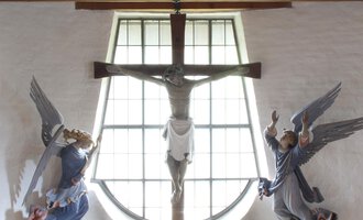 Ein schöner Kirchenraum mit einem Kreuz und Engeln an den Wänden. Der Altar ist mit Blumen geschmückt und hat eine helle, einladende Atmosphäre. | © Ev. Kirche Kleinwalsertal