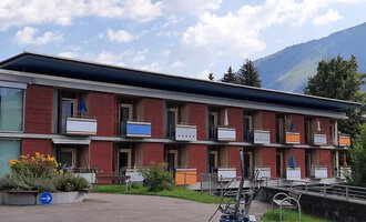 Ein modernes Gebäude mit mehreren Etagen und Balkonen. Im Hintergrund sind grüne Hügel und ein blauer Himmel zu sehen. | © Kleinwalsertal Tourismus | Veronika Senn