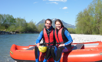 Ein Paar sitzt lächelnd an einem Flussufer in Wasser-Rafting-Ausrüstung. Im Hintergrund sind Bäume und Berge zu sehen. | © MAP-Erlebnis