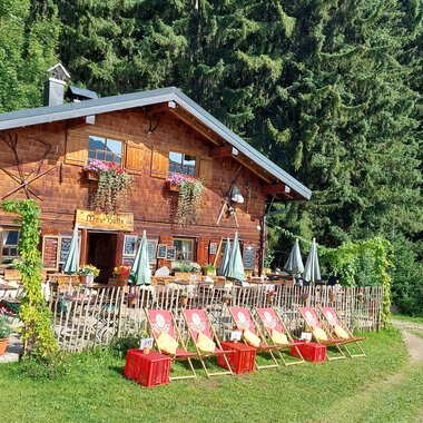 A cozy wooden house surrounded by tall trees. In front of the house, there are chairs and tables in an inviting outdoor area. | © Maxhütte | Viera Krafcik
