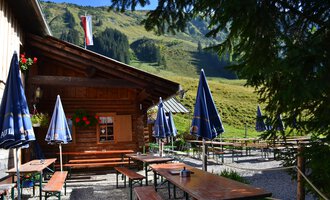 A cozy cabin in the mountains with tables and benches outdoors. In the background, green meadows and mountains can be seen. | © Kleinwalsertal Tourismus