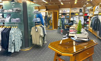 A modern clothing store with various shirts and pants. In the foreground, there is a wooden table, while the shelves in the background are full of clothes. | © Moosbrugger Mode e.U.