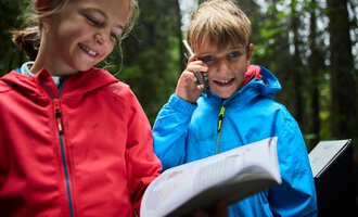 Zwei Kinder stehen im Wald. Das Mädchen lächelt, während der Junge telefoniert und ein Buch hält.