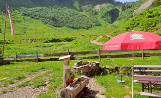 A green mountain landscape with a red sunshade and a fire pit. In the background, gentle hills and forests are visible. | © Kleinwalsertal Tourismus | Steffen Stöhr